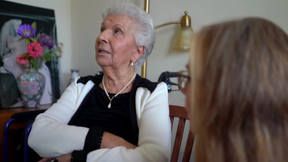 Elderly woman looks off camera and talks to a mature woman with eyes rolling.