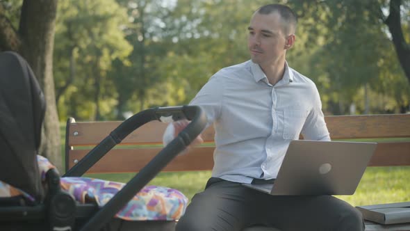 Portrait of Busy Young Businessman Rocking Baby Carriage and Surfing Internet on Laptop Sitting on