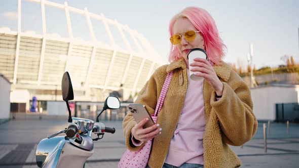 Young Stylish Woman with Pink Hair Drinking Takeout Coffee and Web Surfing on Cellphone Leaning on