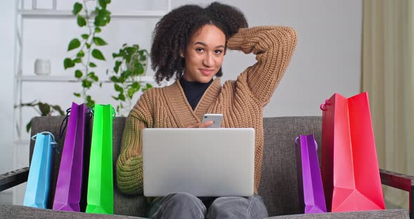 Happy Curlyhaired Ethnic Woman Sitting on Couch with Laptop Looks Into Tap of Mobile Phone Shopping