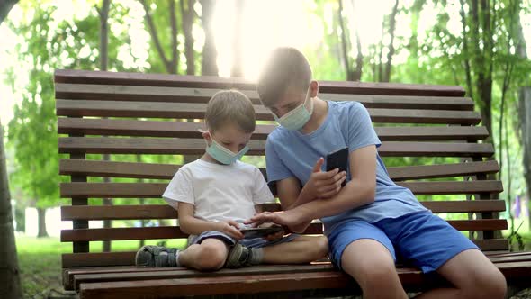 Boys in Medical Masks Play with Smartphones in the Park