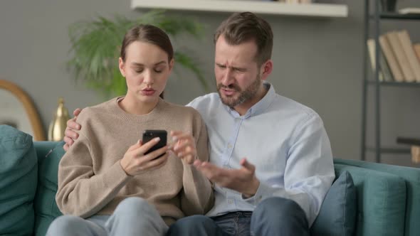 Portrait of Couple Reacting to Loss on Smartphone Sitting on Sofa