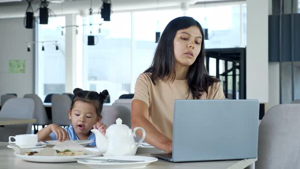 Asian Businesswoman Mom Works Sitting at Table with Tea and Grey Laptop and Child Eats Pizza Slice