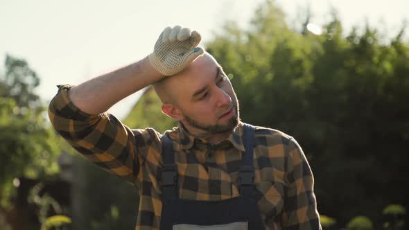 Bearded man in overalls fanning himself with a straw hat, and wiping sweat from his forehead