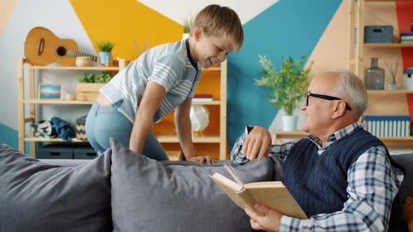 Playful Child and Loving Grandfather Doing High-five While Old Man Reading Book at Home