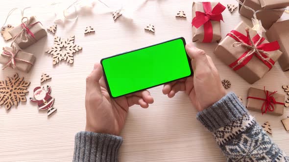 Man holding smartphone with vertical green screen on Christmas background with gifts.