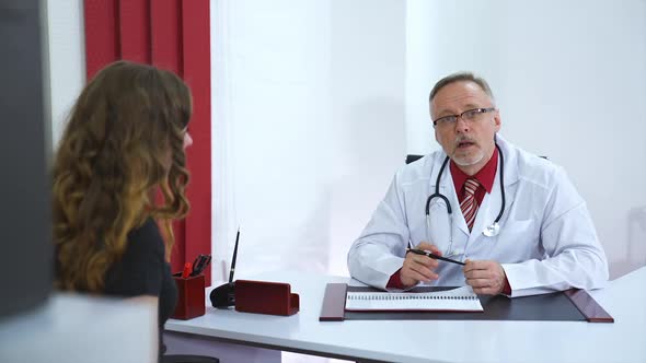 Side view of young female patient on consultation in clinic. Middle aged mature male doctor in white