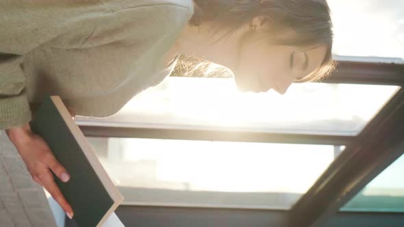 Beautiful Woman Sitting Near Window and Reading a Book on Background of Sunset