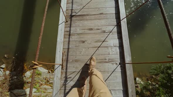Slow Motion POV of Hipster Man Walks on a Wooden Suspended Bridge Over a Mountain River