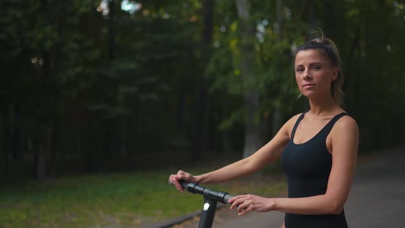 Confident Young Woman Stands Near Her Electric Scooter Wearing Sneakers and a Black Body and Looks