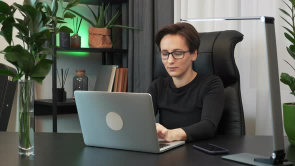 Woman in glasses sitting at modern office workplace working on laptop computer, typing on keyboard