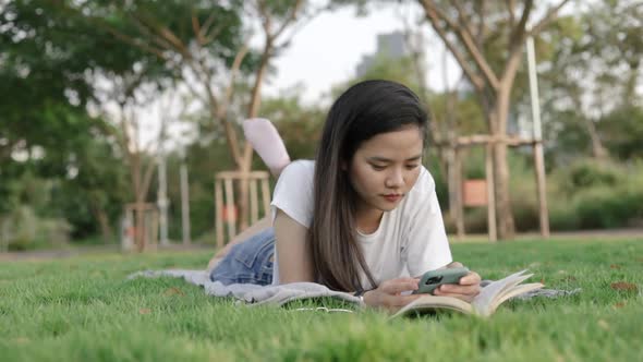 Young Asian woman relaxed reading a book and using a smartphone while lying on the floor grass.