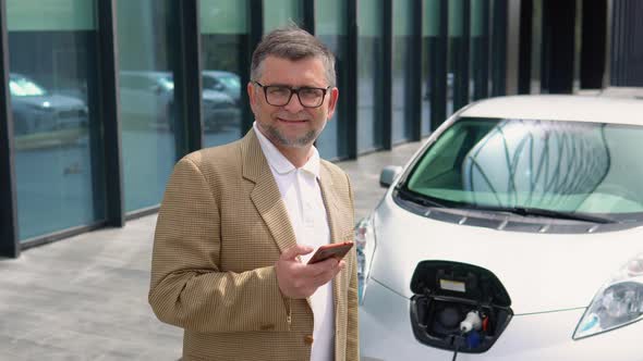 Portrait of Older Man with Smartphone While Charging Electric Car