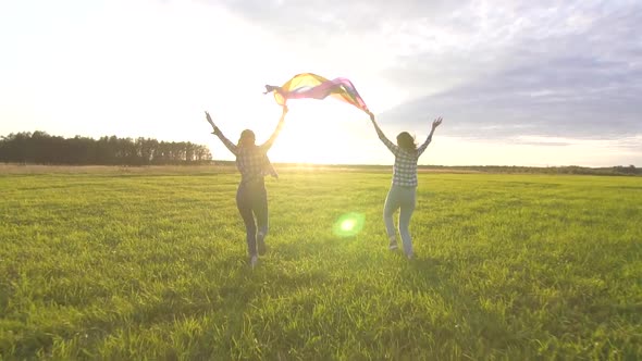 Two Joyful Young Lesbian Girls in Shirts Run Across the Field at Sunset with an LGBT Flag Slow Mo