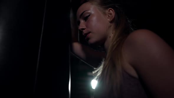 Side View Tired Female Boxer Leaning on Punching Bag Thinking Standing in Darkness with Backlit