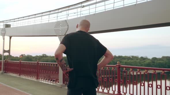 Young White Man in Black Sport Uniform Doing Warmup Neck Before Run Pedestrian Bridge at Dawn