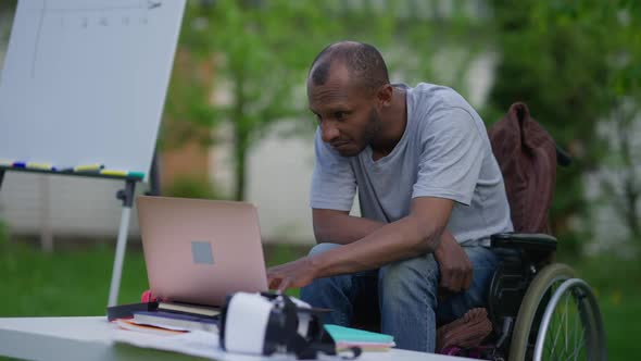 Portrait of Concentrated Handicapped African American Man Surfing Internet on Laptop Sitting in