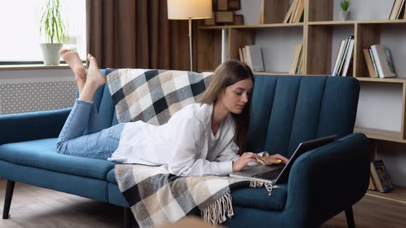 Young Woman Lying on Her Stomach a Sofa Using a Laptop Computer to Surf the Internet with a Happy