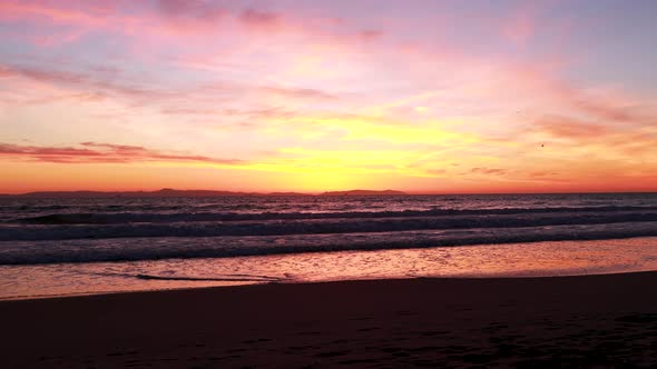 Seagulls fly at the beach during a gorgeous yellow, orange, pink and blue sunset with the Huntington