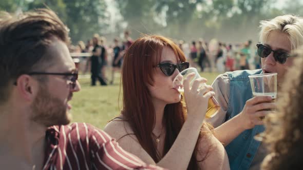 Group of friends sitting on grass together at music festival and drinking beer while chatting. Shot