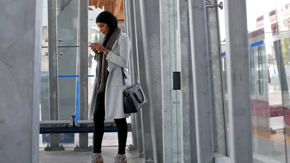 Side view of young Asian woman in hijab using mobile phone at railway station 4k