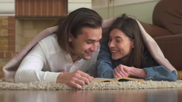 Joyful Couple in Love Lying on the Carpet in the Bedroom Covered with a Blanket Reading a Book