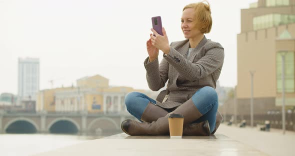 40Yearold Beautiful Woman Sits on the Embankment and Takes a Photo of the Cityscape with Her