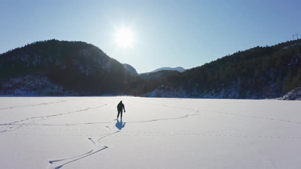 Aerial following man silhouette in bright sun, ice skating on frozen lake Mountains Background