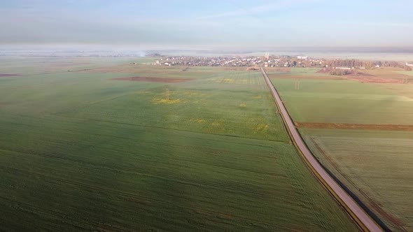 Drone flying over a small country road with green farming fields in the early misty morning with sun