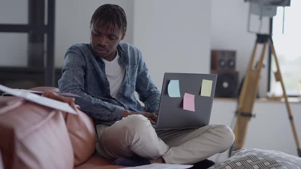 Busy Young Man Looking Through Paperwork Typing on Laptop Keyboard Sitting on Comfortable Couch in