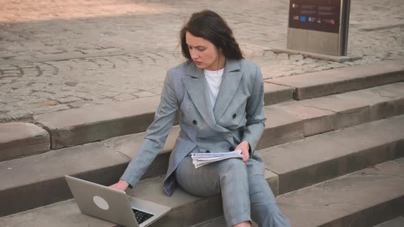 Upset Young Woman Sitting on Stairs While Using Laptop Working with Papers Documents on the Street