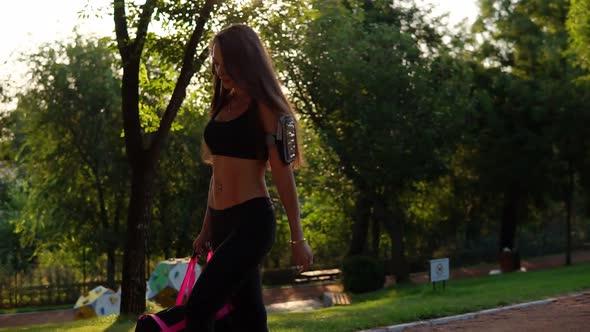 Beautiful Brunette Woman in Short Sport Top and Armband Walking with Sport Bag in the Park