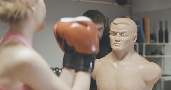 Back View of Blond Female Boxer Punching Mannequin with Blurred Personal Trainer Talking at the alt