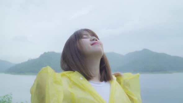 Young Asian woman feeling happy playing rain while wearing raincoat standing near lake.