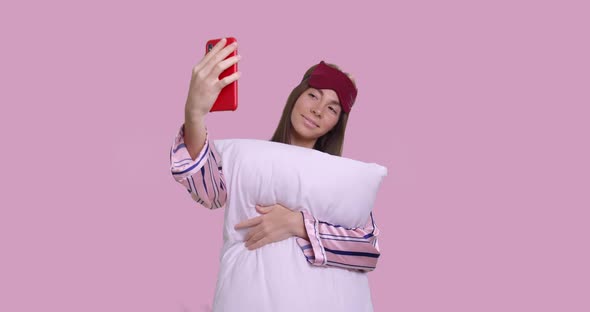 Pretty Female Makes Selfie in Pajamas and Pillow Isolated Over Pink Background