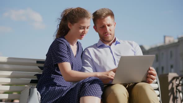 Happy Young Couple Using Laptop Computer Sitting on Bench in City Outdoor