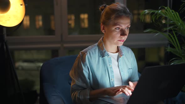 Woman is Sitting in the Armchair and Working on a Laptop at Night or Texting Someone