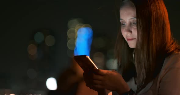 At Night, a Young Girl Holds a Smartphone in Her Hands and Looks at the Screen