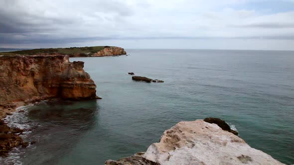 A long pan of a rocky coast into a cliff made of orange and red rocks layered from years of erosion.