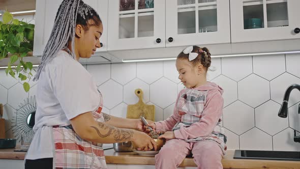 Active Little African American Girl Whisking Dough in Bowl Her Mother Watching Her at Kitchen Both