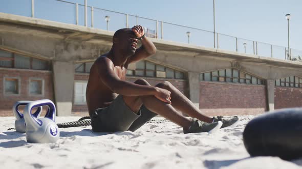 Tired african american man sitting, wiping his forehead, taking break in exercise outdoors on beach