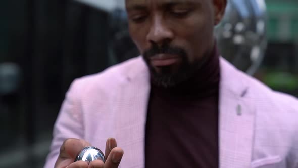 Cool Black Man Is Holding and Rotating Metal Balls in Hand, Looking at Camera Enigmatically