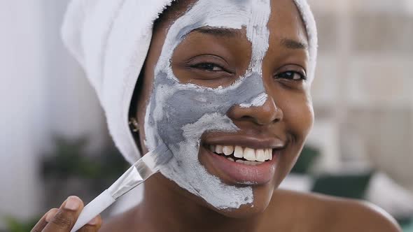 Smiling Afro-American Girl with Towel on the Head Applying a Cleansing Clay Mask on Face