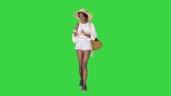 Pretty African American Woman in a Straw Hat Texting on Her Phone While Walking on a Green Screen