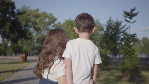 Back View of Older Sister Spending Time with Younger Brother Chatting in the Summer Park