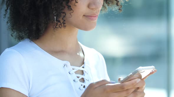 Attractive African American Girl Smiling Cutely, Holding Smartphone in Hands, Typing Message on