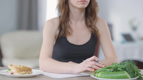 Healthy and Unhealthy Food on Table with Blurred Unrecognizable Slim Young Woman Writing Calories