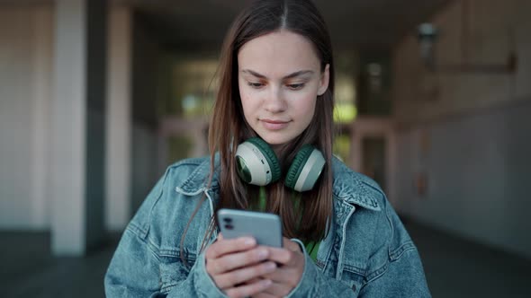 Positive blonde woman in headphones wearing jeans jacket typing by phone