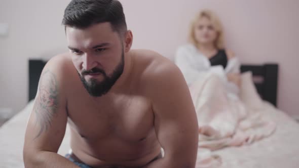 Close-up of Angry Caucasian Man Sitting on Bed. Blurred Blond Woman Lying at the Background. Wife