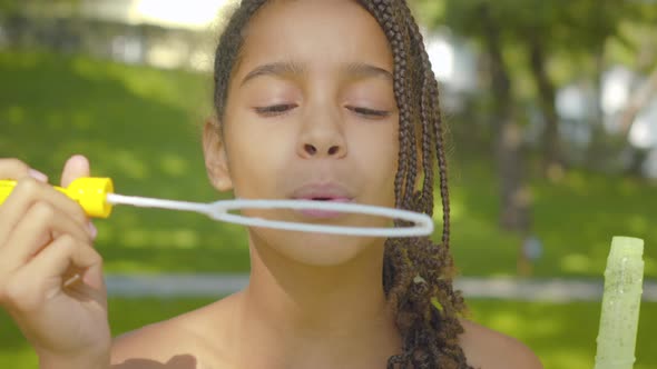 Close-up of Charming Happy African American Girl Blowing Soap Bubbles in Sunny Summer Park. Portrait